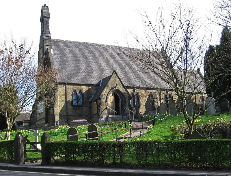 St Paul's Church, a grey stone Victorian church with a slate roof and a tall bell turret, seen from the road behind a low hedge, with gravestones and daffodils in the churchyard and bare spring trees.