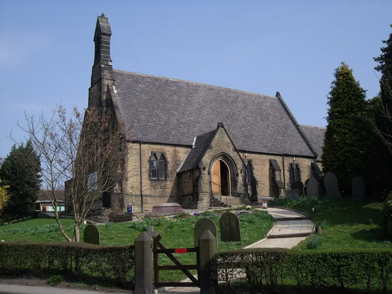 St Paul's Church in Hasland from the roadside: a stone church with a pitched slate roof, a pointed bell turret and an arched porch, behind a hedge and gate, with bare trees and gravestones.