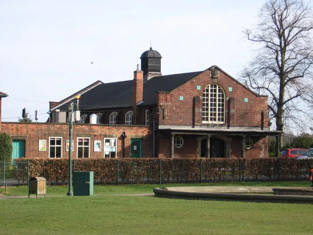 Hasland Village Hall, a red-brick hall with a large arched window, a small cupola on the roof and a colonnaded porch, seen across the lawn of Eastwood Park with a bare tree beside it.