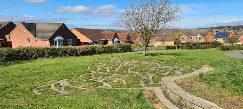 A grassy open space on the Hasland Green estate with a large path map made of brick, stone and slate set into the lawn, a bare tree, hedges and modern red-brick houses under a blue sky.
